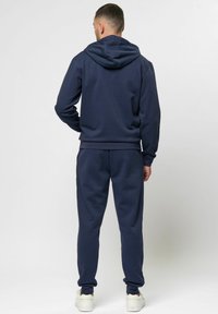 Tumed sinine kapuutsiga ja looderdurve komplekt pehmest kangast. Omab kapuutsi, soonikkoesid varrukates ja mugavat lõiget, koos valgete tossudega.