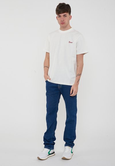 T-shirt bianco in cotone con testo ricamato rosso sul petto, abbinato a jeans di denim blu e sneakers bianche con dettagli verdi.