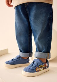 Baskets en toile bleue avec semelles blanches plates, ornées d'imprimés de personnages de dessins animés sur les côtés. Portées avec des pantalons en denim retroussés.