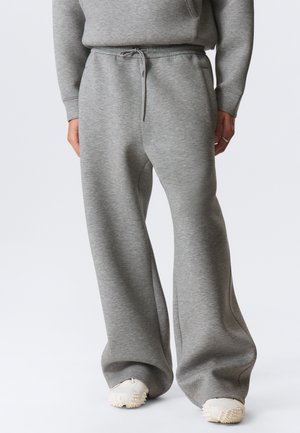 Henkilö, joka käyttää väljät harmaat collegehousut, joissa on kiristysnauha vyötäröllä, ja valkoiset, kuvioidulla pohjalla varustetut tennarit, seisomassa valkoisen taustan edessä.