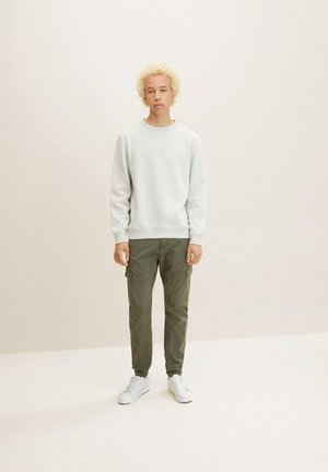 Sudadera de cuello redondo gris claro combinada con pantalones cargo verde oliva que tienen múltiples bolsillos. Llevada con zapatillas blancas sobre un fondo liso.