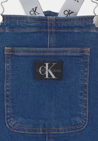 Džínové laclové kalhoty s nastavitelnými šedými popruhy, které mají černé logo "CK", tmavě modrou látku, kontrastní oranžové švy a detail zadní kapsy.