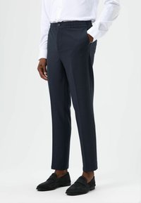 Dunkelgraue, maßgeschneiderte Hosen mit Knopf und elastischem Bund, ausgestattet mit Seitentaschen und geradem Bein. Getragen mit schwarzen Loafers.