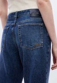 Σκούρο μπλε τζιν με ψηλή μέση, εμφανείς πίσω τσέπες. Διαθέτει δερμάτινο ένθετο με ραφή και μικρό κεντημένο λογότυπο κοντά στην τσέπη.