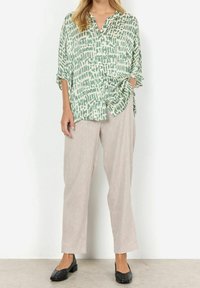 Chemise boutonnée à motifs verts et blancs à manches courtes, associée à un pantalon beige ample et des chaussures plates noires.