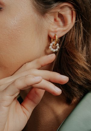 Goldener Creolen-Ohrring mit kleinen weißen Perlen, die ein kreisförmiges Design bilden. Nahaufnahme des Ohrrings, getragen im Ohr, mit Fingern, die die Kieferlinie berühren.