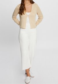 Beigevärinen ribbineulottu neuletakki pyöreällä pääntiellä ja napitusyksityiskohdalla, yhdistetty valkoisiin leveälahkeisiin housuihin ja samansävyisiin liukusandaaleihin. Sileä pinta.