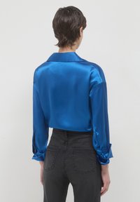 Persona con capelli corti castani, vestita con una blusa lucida blu a maniche lunghe e jeans neri a vita alta, mostrata di spalle.
