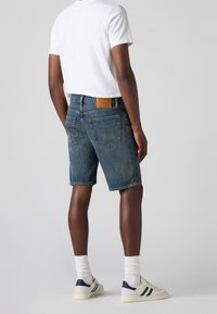 Shorts en denim de couleur bleu moyen, avec une finition délavée, cinq poches et un patch en cuir au dos. Portés avec un t-shirt blanc uni et des baskets.