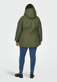 Olivgrön, huva parka jacka med åtdragen midja, tillverkad av ett mjukt, syntetiskt material och långa ärmar, matchad med blå denimjeans.