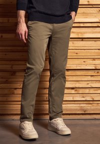 Khaki chinos, joissa on slim fit -malli, sileä pinta ja viisi taskua, yhdistettyinä valkoisiin tennareihin. Taustalla puupaneeli.