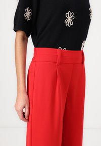 Pantalons larges rouges à taille haute avec des plis. Tissu lisse avec une finition soignée, complété par un haut noir orné de broderies florales.