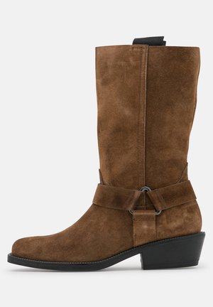 Botte mi-mollet en daim marron avec talon bas noir, sangle décorative avec anneau en métal, et une languette noire en haut.