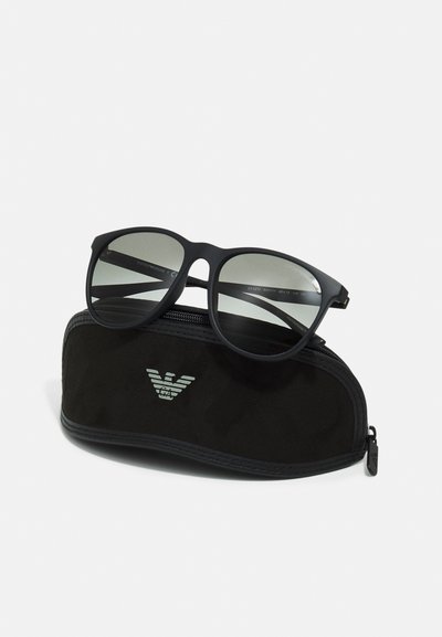 Occhiali da sole neri con lenti scure poggiano su una custodia nera morbida. La custodia presenta un logo argentato e ha una chiusura con zip.