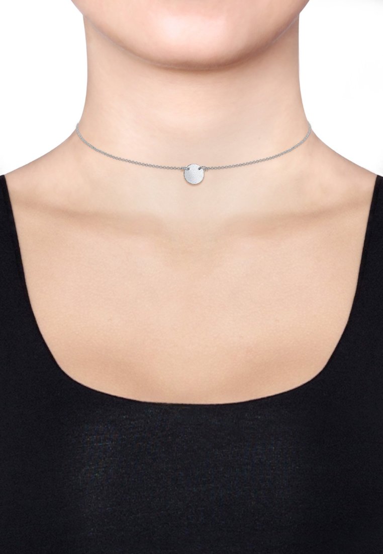 Sølv choker halskæde med en tekstureret cirkulær vedhæng, der har en fin kæde og glat overflade, iført over en mørk top.