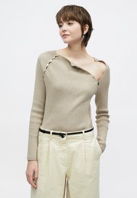Bež pulover z rebrastim vzorcem in asimetričnim izrezom z gumbi na enem ramenu. Kombiniran z visokim pasom kremnih hlač in črnim pasom.