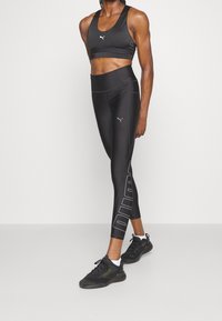 Czarny sportowy crop top z wycięciem na plecach i detalem z logo, zestawiony z czarnymi legginsami o wysokim stanie z wyraźnym wzorem marki.