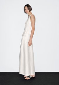 Vit, ärmlös maxiklänning med en åtsittande bodice och ett flödande kjol, tillverkad av texturerat tyg. Kombinerad med svarta, remmade sandaler.