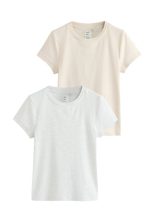 Ung kvinde med brunt hår bærer en kortærmet, off-white top og lyse stribede bukser, står mod en ensfarvet lys baggrund.