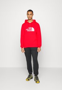 Sweatshirt com capuz vermelho com logótipo branco, combinado com calças de fato de treino pretas e sapatos de caminhada cinza-esverdeados com detalhes amarelos.