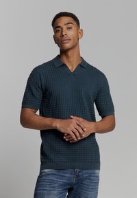 Gebreide polo met korte mouwen in donker te groen met een gestructureerd patroon, V-hals en gebreide kraag en manchetten. Draagt met blauwe jeans.