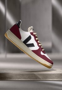 Sko med hvitt lær på overdelen, maroon semsket skinn detaljer, svart logo, beige mellomsåle og såle av gummi.