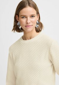 Pull crème avec un motif de tricot en losange texturé et un col rond. De grandes boucles d'oreilles géométriques en argent accentuent le look.