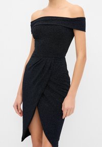 Femme portant une robe noire ajustée à épaules dénudées avec un tissu texturé et une fente à l'avant, debout devant un fond blanc uni.