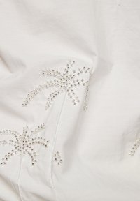 White fabric featuring intricate beadwork forming palm tree designs. Beads are clear and metallic, adding texture and detail to the material.