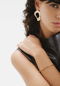 Pendientes grandes con textura dorada y una pulsera de eslabones dorados, llevados con un top de cuero negro, mostrando piel suave y luz natural.