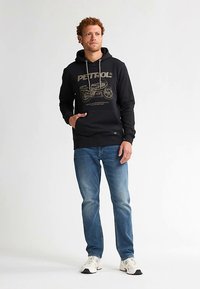 Čierna mikina s kapucňou s grafikou "PETROL" v zlatej farbe a motocyklovým dizajnom, kombinovaná s modrými džínsami a bielymi teniskami; obsahuje kapucňu so šnúrkou a predné vrecko.