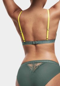 Die Rückansicht einer Frau, die grüne Netzspitzenunterwäsche mit gelben BH-Trägern trägt, vor einem schlichten weißen Hintergrund.