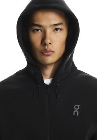 Musta vetoketjullinen huppari, joka on valmistettu pehmeästä kankaasta, siinä on etuvetoketju, säädettävä nyörillinen huppu ja pieni harmaa logo rinnassa.