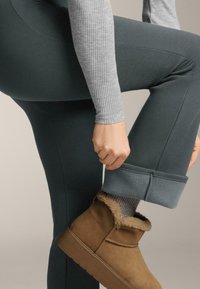 Tmavě zelené žebrované legíny jsou zkombinovány s béžovými kotníkovými botami s kožešinovým podšíváním. Na horní části ruky je vidět šedý dlouhý rukáv.