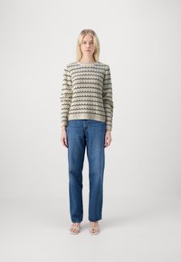 Maglione a maglia colore crema con motivi ondulati neri, collo rotondo e maniche lunghe, abbinato a jeans blu dritti e sandali chiari.