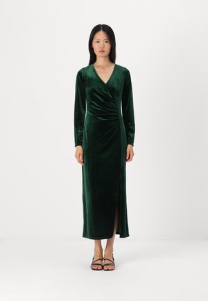 Samtgrünes Kleid mit langen Ärmeln, tiefem V-Ausschnitt, seitlichem Schlitz und Raffungen an der Taille. Kombiniert mit schwarzen, schnürbaren Sandalen.