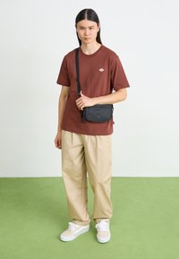 Kleine zwarte crossbody tas met een ritssluiting en een verstelbare band, gemaakt van gladde stof, vastgehouden door een persoon in een bruine t-shirt en beige broek.