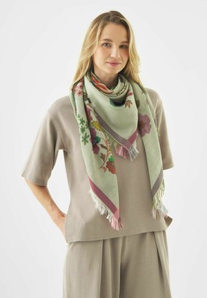 Jonge vrouw met blond haar, gekleed in een beige top met korte mouwen en een broek, met een groene sjaal met bloemenpatroon en franjes omgeslagen.