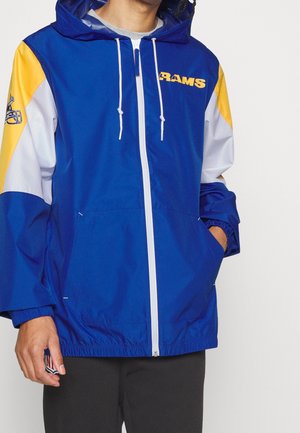 Personne portant une veste à capuche zippée Rams bleue, blanche et jaune avec cordons et main dans la poche, cadrée au niveau du cou et des cuisses.