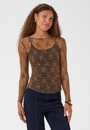 Jeune femme aux cheveux bouclés portant un haut marron transparent à manches longues par-dessus un débardeur marron et un jean bleu foncé, debout devant un fond uni.