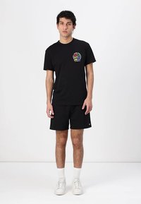 Svart bomull T-skjorte med flerfarget brodert logo på brystet, sammen med svarte shorts. Modellen har på seg hvite joggesko og ankelsokker.