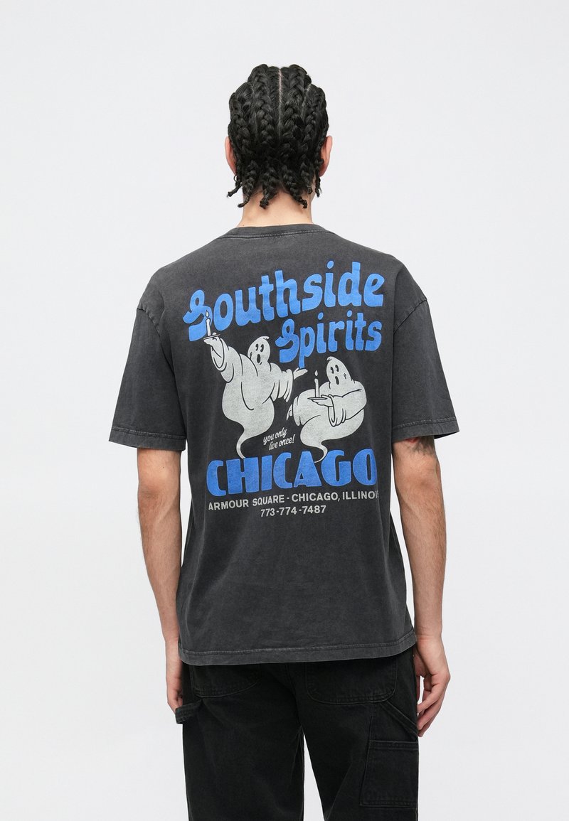 Melnas kokvilnas T-krekls ar brīvu piegriezumu. Uz muguras ir zilā un pelēkā krāsā drukāts grafisks attēls ar diviem karikatūriskiem spokiem un tekstu.