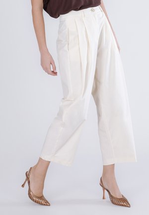 Personne portant un pantalon blanc taille haute à jambes larges avec plis, associé à des escarpins slingback métalliques bronze, sur un fond clair.