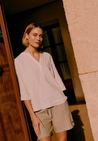 Woman with short blonde hair stands in a doorway wearing a white top and beige shorts, lit by warm sunlight.