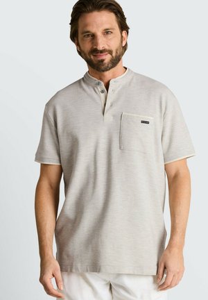 Mann mit Bart, der ein hellgraues Henley-Shirt mit kurzen Ärmeln und Brusttasche sowie weiße Hose vor einfarbigem Hintergrund trägt.