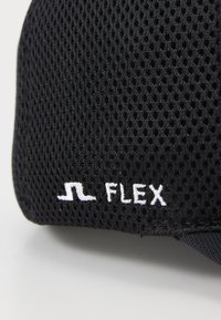 Melnas acu cepures ar izteiktu balto izšūto logotipu "JL FLEX" sānos. Elpojoša tekstūra ar strukturētu dizainu.