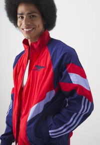 Crvena i mornarsko plava jakna na vjetar s horizontalnim prugama i izvezenim logotipom. Ima zatvarač sprijeda i elastične manžete. Lagani materijal.