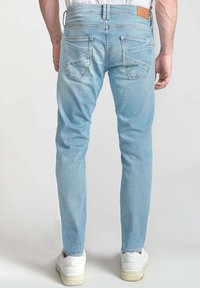 Jean skinny bleu clair avec un effet délavé, cinq poches et un patch en cuir beige au niveau de la taille. Porté avec des baskets blanches.