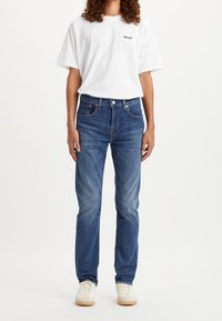 Personne portant un t-shirt blanc Levi's, un jean droit classique bleu et des baskets blanches, debout devant un fond blanc uni.