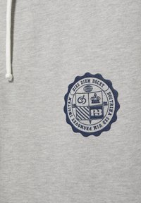 Hellgrauer Stoff mit dunkelblauem rundem Emblem mit lateinischem Text und grafischen Symbolen neben einem weißen Kordelzug.